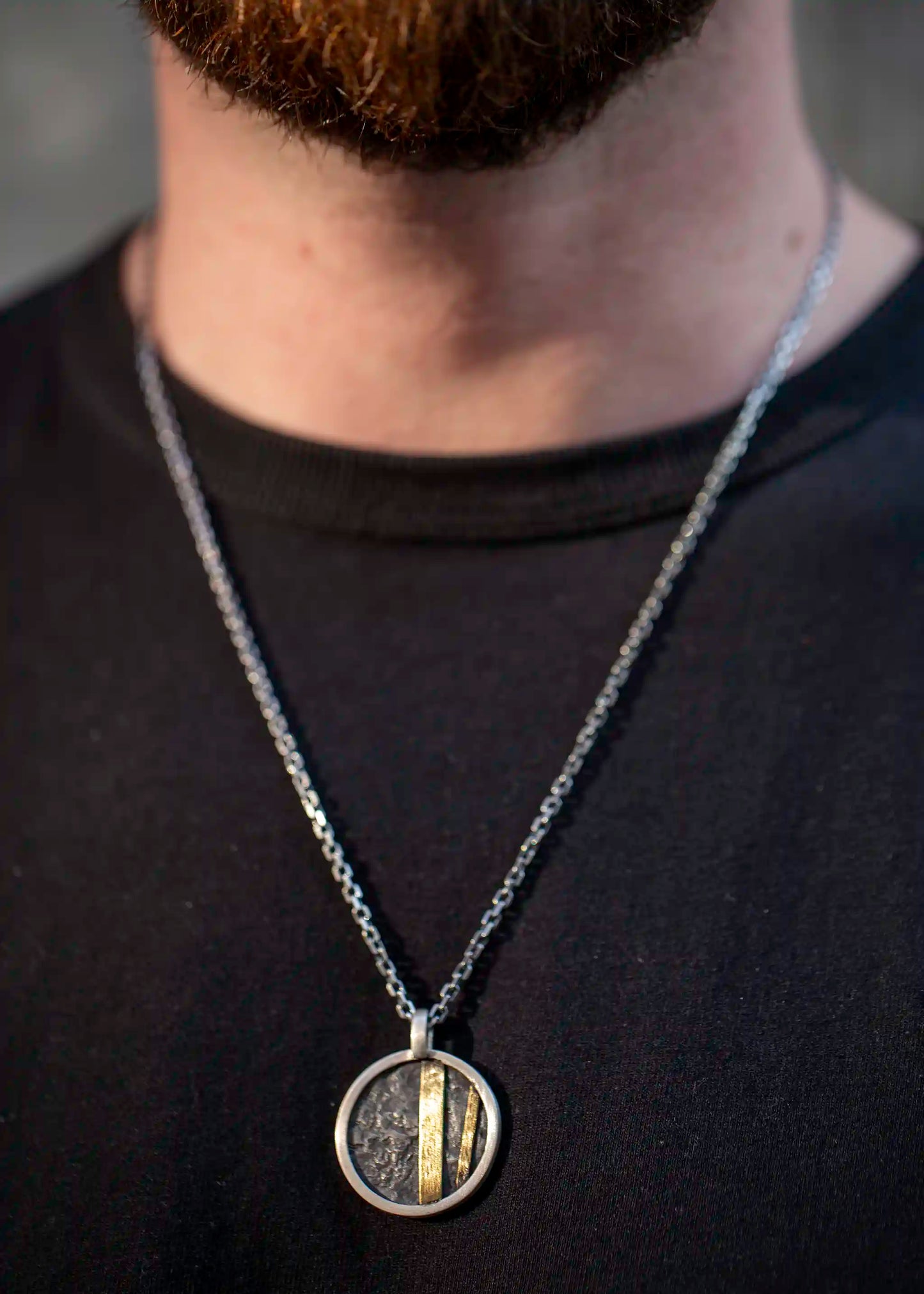 Person wearing the 42 Pendant—round silver with textured dark surface and brass inlays—on a silver chain.