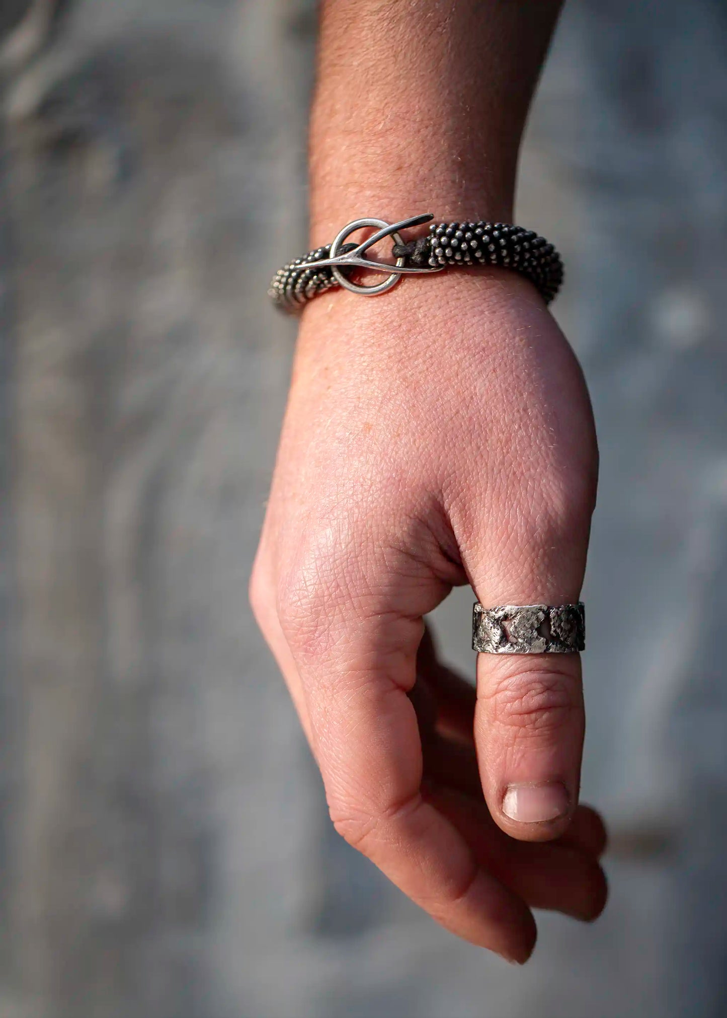 Textured silver beaded bracelet with a hook closure, worn on a wrist with a rugged silver ring, against a blurred background.