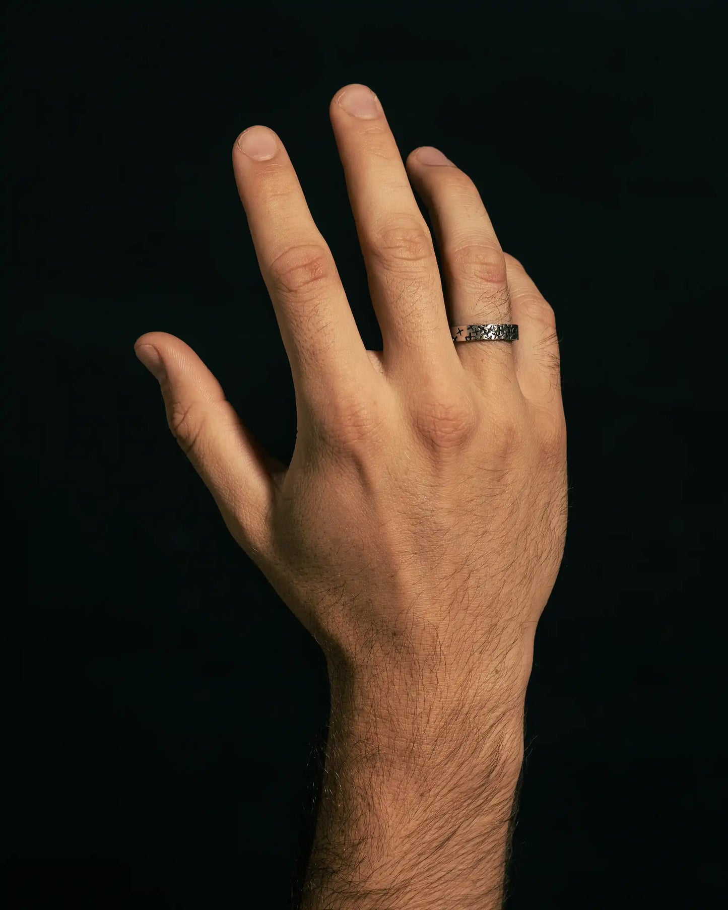 Hand wearing a silver ring with bold black cross patterns, set against a dark background.