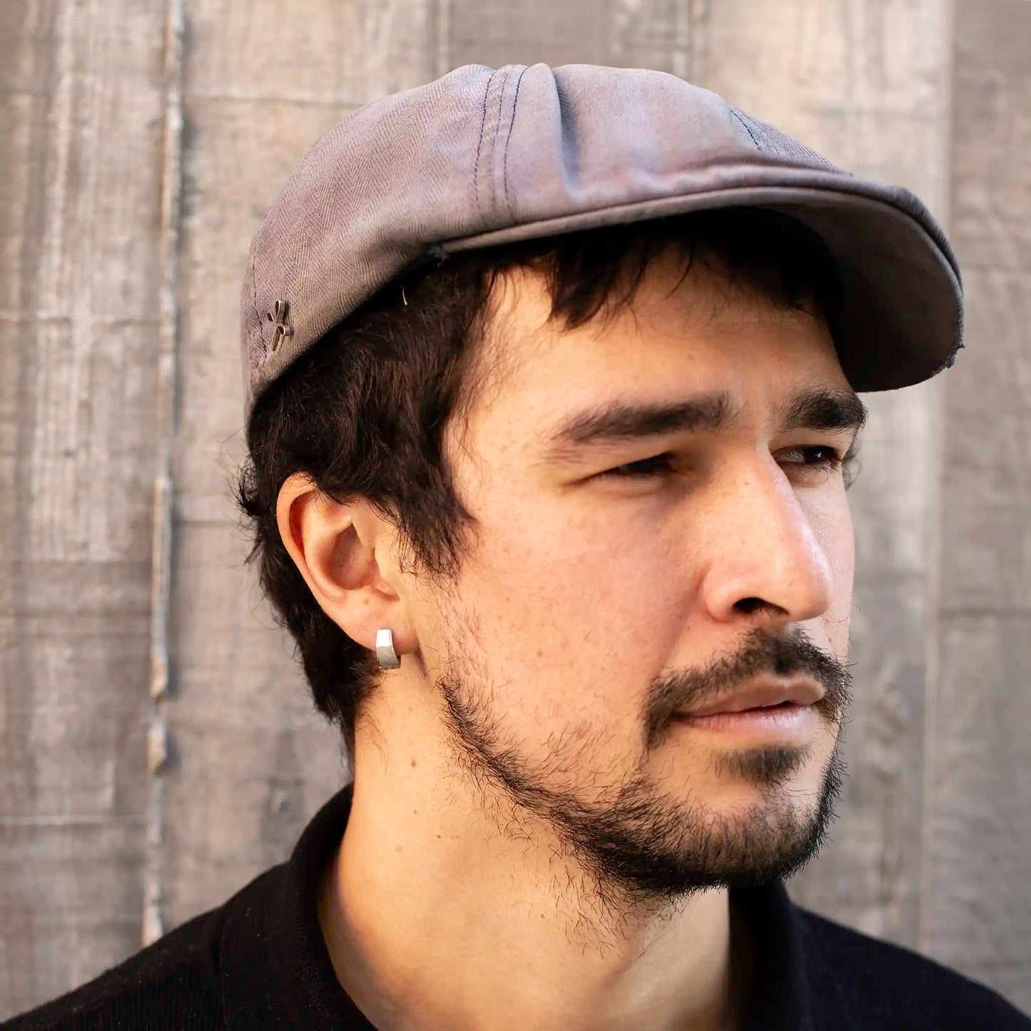 Man with dark hair and beard wearing a gray flat cap, black collared shirt, and Ekko Earrings, standing against a wooden wall with sunlight on his face.