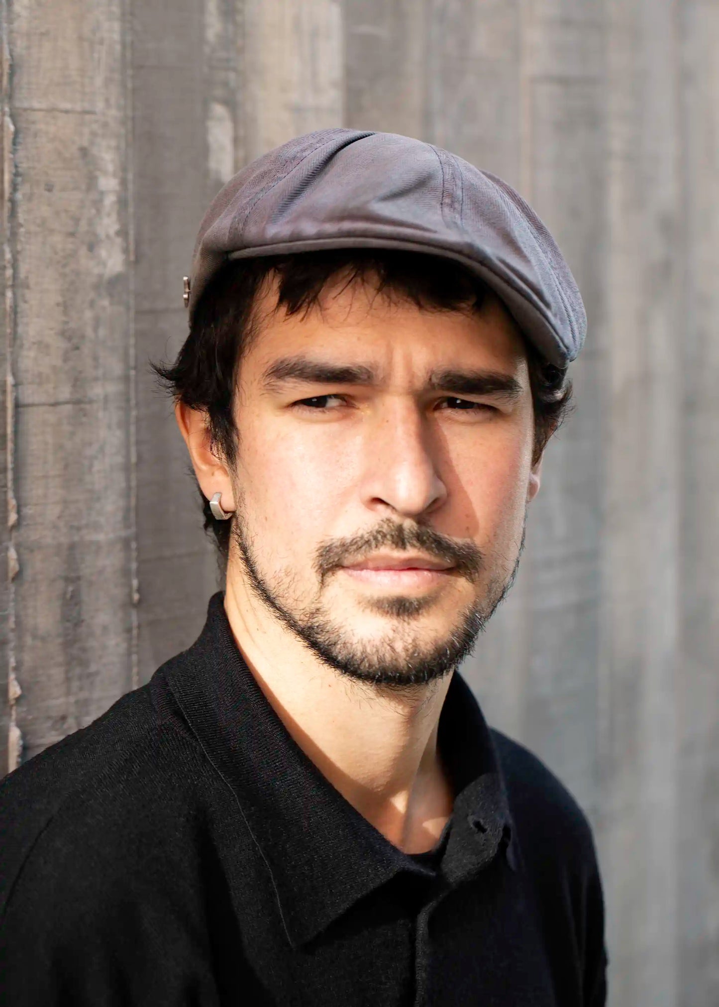 Man with dark hair and beard wearing a gray cap, black collared shirt, and Ekko Earrings, standing against a textured wall with sunlight on his face.