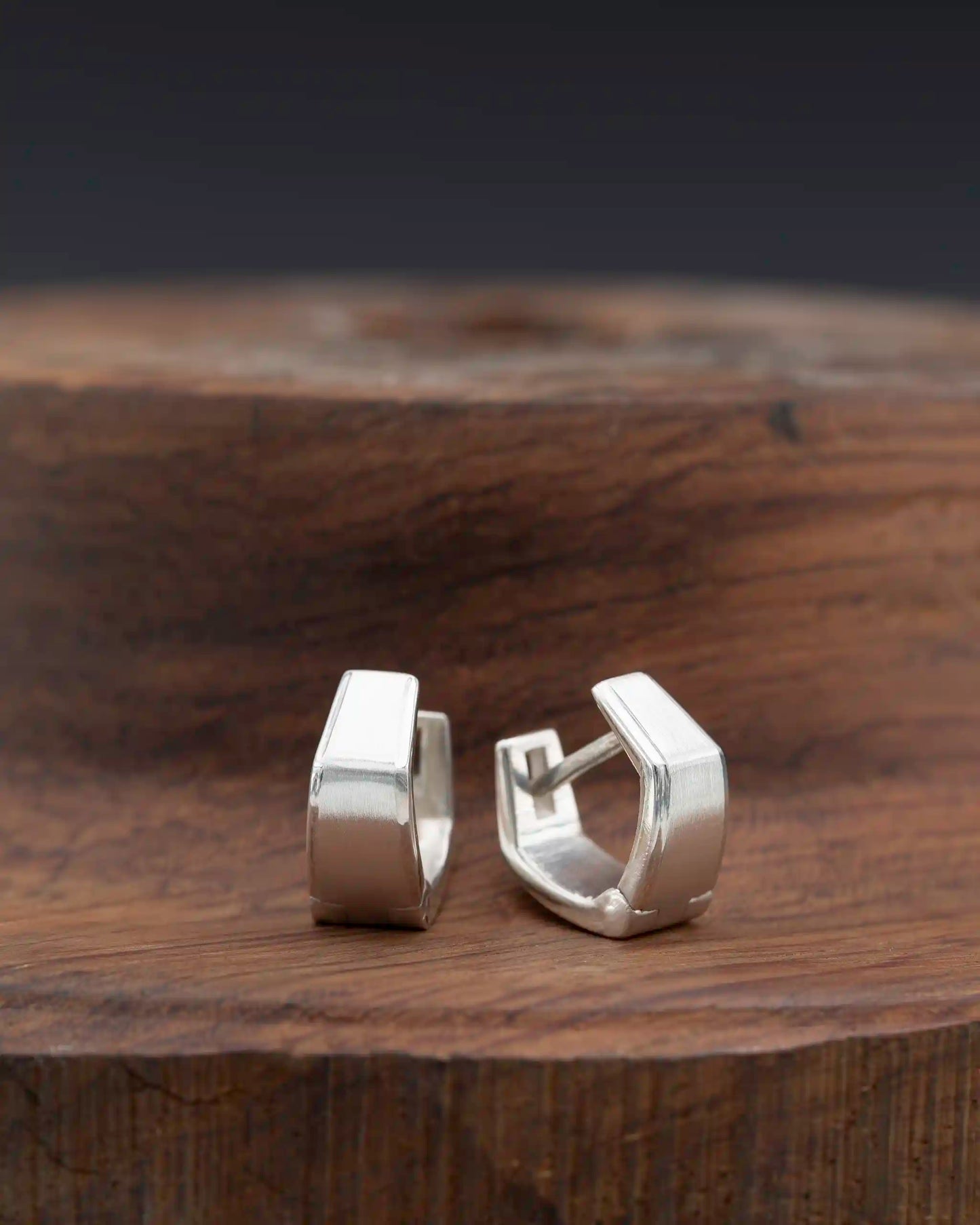 Modern minimalist silver huggie earrings with geometric design, displayed on a rustic wooden surface.