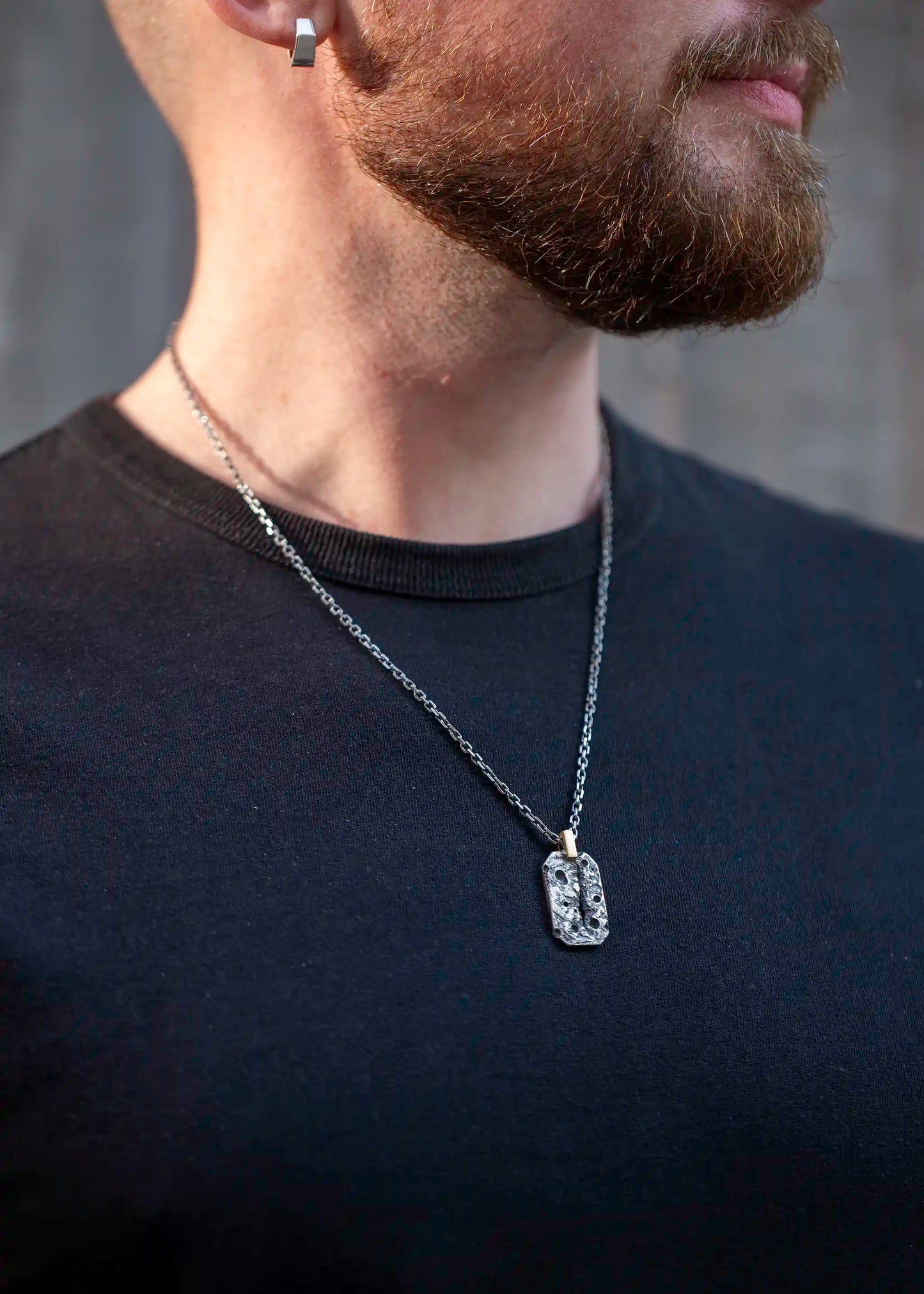 Rugged textured silver pendant with black stone details on a silver chain, worn by a bearded man in a black shirt.