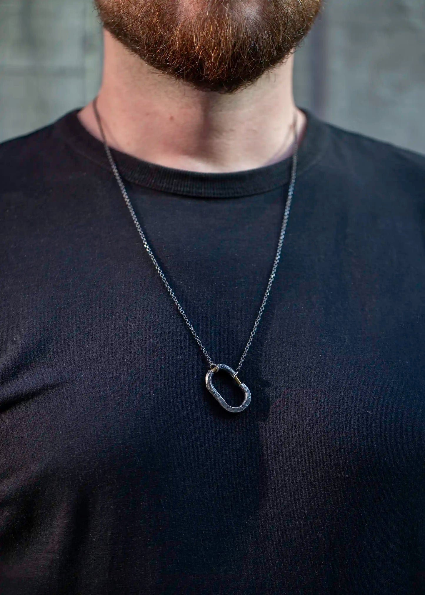 Man in a black shirt wearing a silver chain necklace with a textured oval pendant and brass detail.