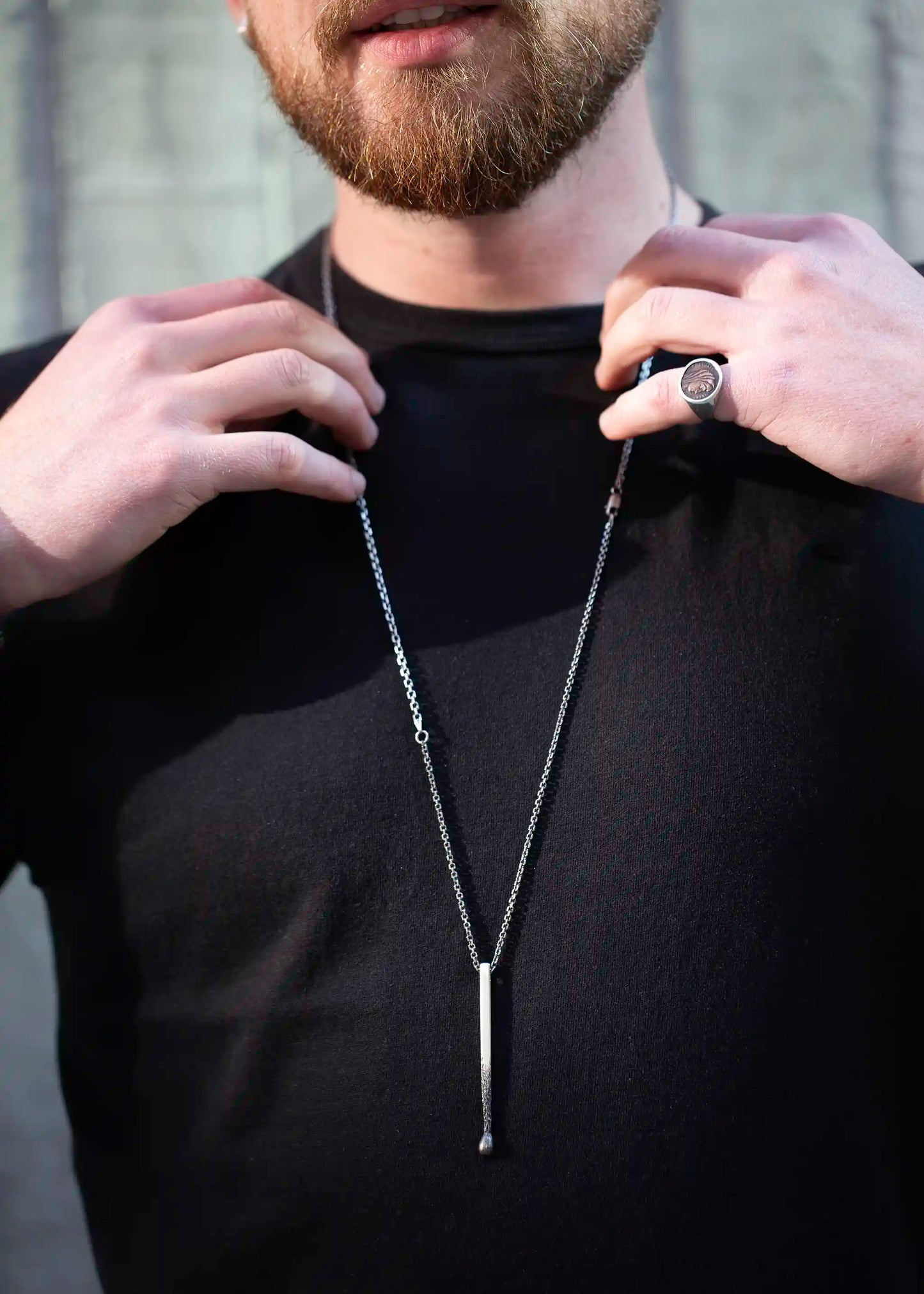 Man in black shirt adjusting a silver necklace with vertical bar pendant, wearing a silver signet ring.