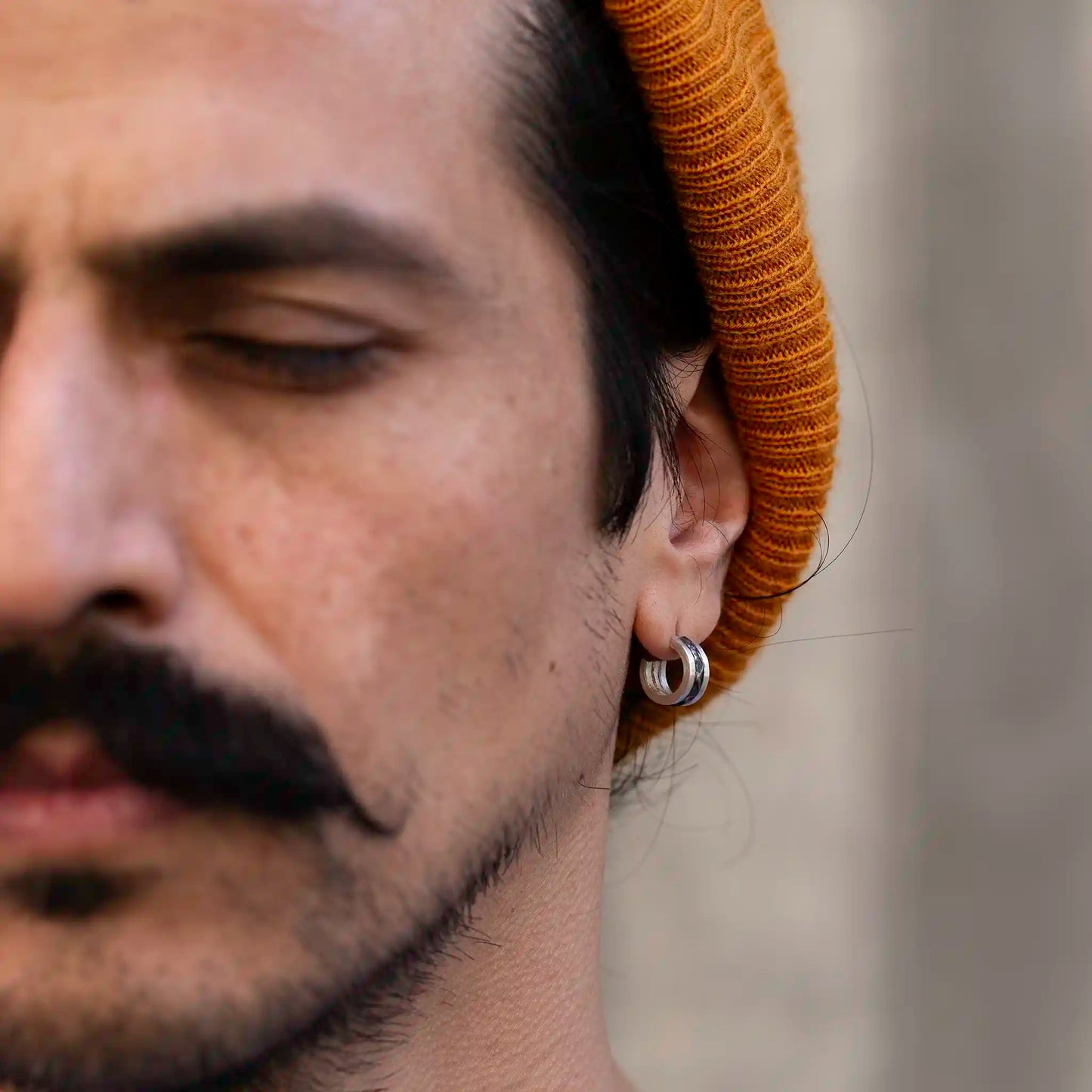 Close-up of a man with a mustache wearing a mustard beanie and a silver Mountain earring, eyes closed, blurred background.