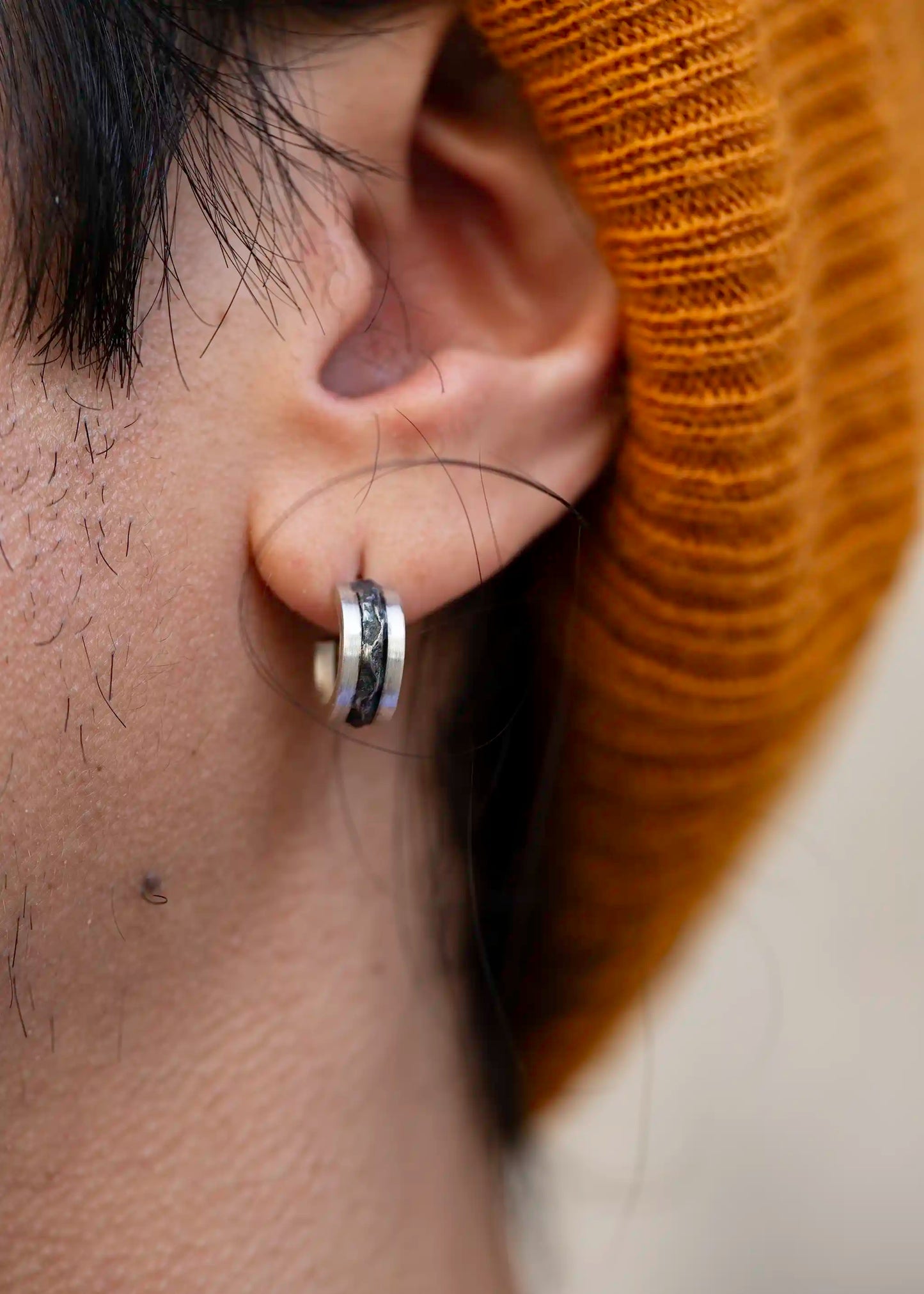Close-up of a man’s ear with a silver Mountain earring featuring a black textured design, mustard beanie in background.