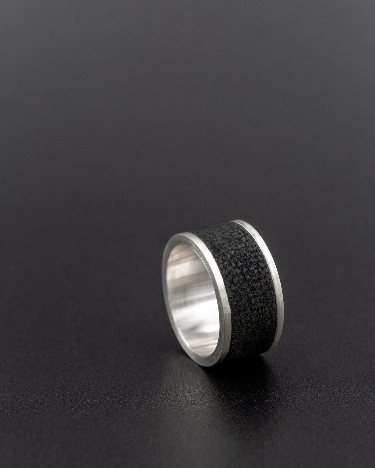 Silver ring with a wide black textured leather inlay, photographed on a dark background for a sleek, modern look.