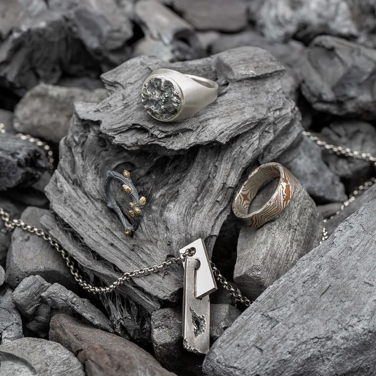 Display of handcrafted jewelry including silver and mixed-metal rings, a pendant necklace, and an oxidized gold-accented piece, arranged on rugged wood and stones.