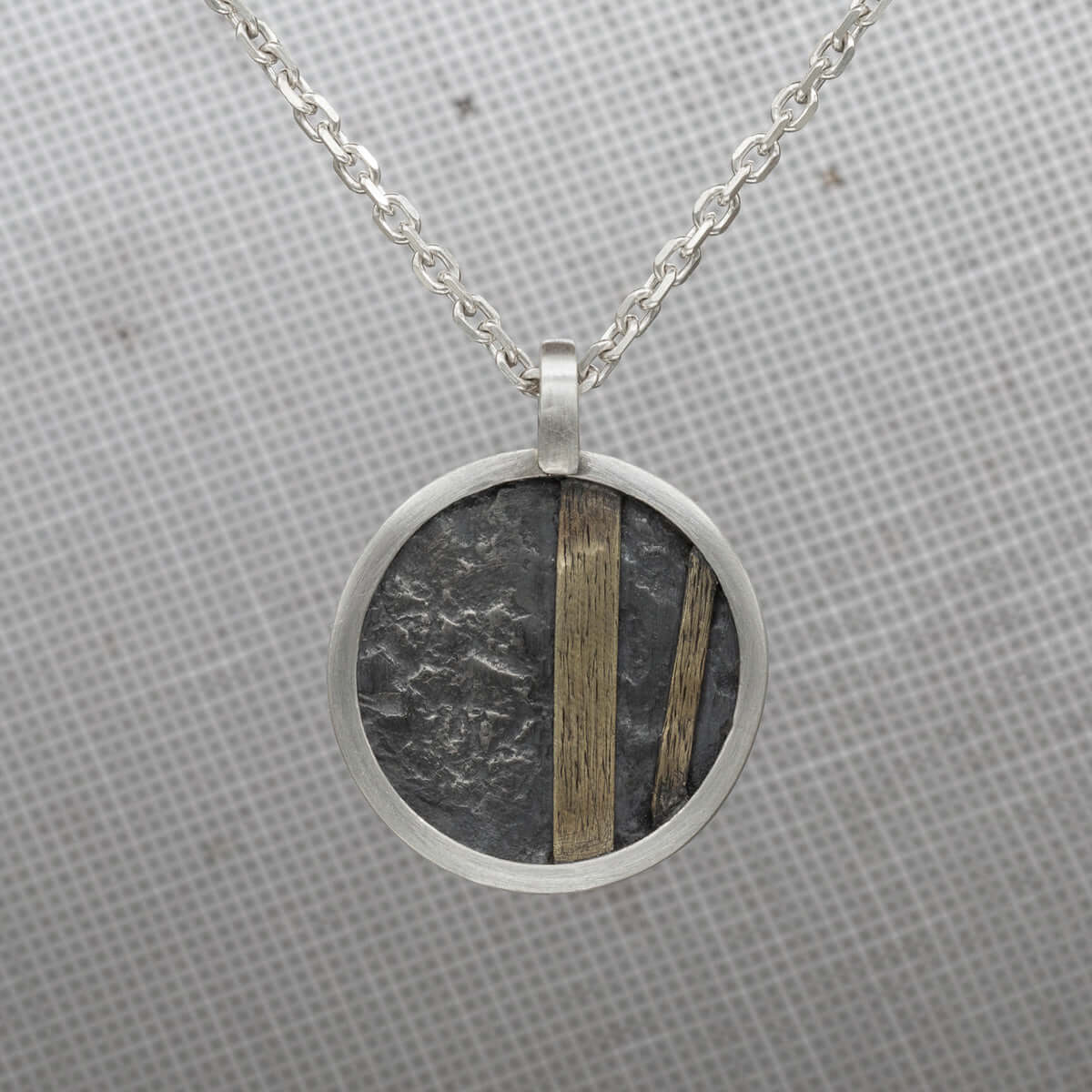 Close-up of a handcrafted round pendant featuring oxidized silver with textured detailing and gold accents, hanging on a delicate silver chain against a subtle patterned background.