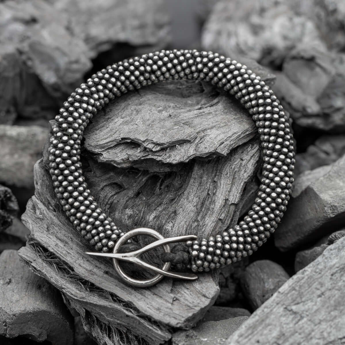 Close-up of a textured silver bracelet with intricate beaded details and a wishbone-style clasp, placed on rugged, natural wood and stone background.