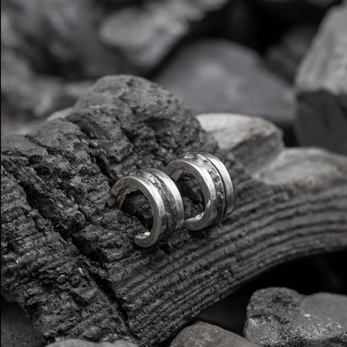 Close-up of two handcrafted silver earrings with a rugged, textured design placed on a piece of charred wood and surrounded by dark stones, creating a raw, natural aesthetic.